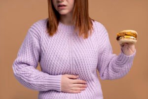 Mujer con malestar abdominal sosteniendo una hamburguesa, representando el impacto de la alimentación alta en grasas y ultraprocesados en el desarrollo del hígado graso no alcohólico